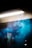 A fluorescent light fixture is mounted on a surface with blue smoke swirling beneath it, creating a mysterious and atmospheric visual effect. The background is mostly dark, allowing the smoke's color to stand out.