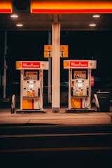 A pair of gas pumps stand under an illuminated canopy, with vibrant red and yellow colors. The pumps belong to a Shell gas station, displaying the V-Power Nitro+ branding. The setting is dimly lit, suggesting nighttime or evening.