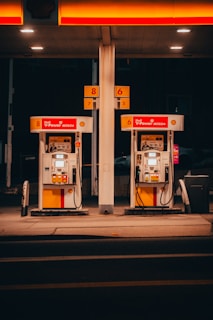 A pair of gas pumps stand under an illuminated canopy, with vibrant red and yellow colors. The pumps belong to a Shell gas station, displaying the V-Power Nitro+ branding. The setting is dimly lit, suggesting nighttime or evening.