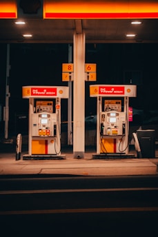 A pair of gas pumps stand under an illuminated canopy, with vibrant red and yellow colors. The pumps belong to a Shell gas station, displaying the V-Power Nitro+ branding. The setting is dimly lit, suggesting nighttime or evening.