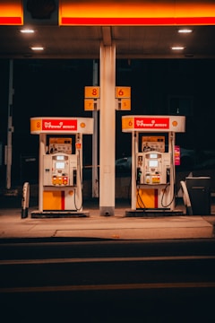 A pair of gas pumps stand under an illuminated canopy, with vibrant red and yellow colors. The pumps belong to a Shell gas station, displaying the V-Power Nitro+ branding. The setting is dimly lit, suggesting nighttime or evening.