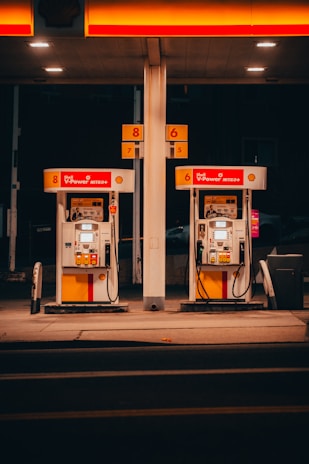 A pair of gas pumps stand under an illuminated canopy, with vibrant red and yellow colors. The pumps belong to a Shell gas station, displaying the V-Power Nitro+ branding. The setting is dimly lit, suggesting nighttime or evening.