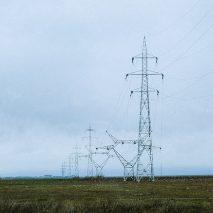A series of tall electrical pylons stretch across a grassy, open landscape under a cloudy sky. The structures are composed of intricate metal frameworks supporting electrical wires.