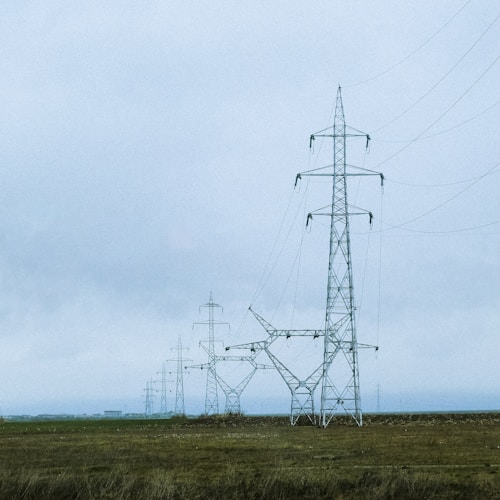 A series of tall electrical pylons stretch across a grassy, open landscape under a cloudy sky. The structures are composed of intricate metal frameworks supporting electrical wires.