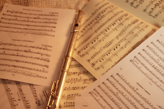 A silver flute is placed among several sheets of musical notation. The sheets feature complex arrangements and notes, spread out on a neutral background, suggesting a focus on music and practice.