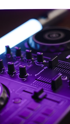 A close-up view of a DJ mixer with various control knobs and sliders. The equipment features intricate details highlighted by a purple hue and a distinct light source. The brand name 'Pioneer DJ' is visible on the surface.