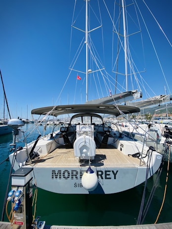 A sailboat docked at a marina, featuring a spacious deck and dual steering wheels. The sky is clear with bright blue hues, and several other boats are visible in the background. A small red flag can be seen on the boat's mast, and the marina is bustling with nautical activity.