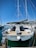 A sailboat docked at a marina, featuring a spacious deck and dual steering wheels. The sky is clear with bright blue hues, and several other boats are visible in the background. A small red flag can be seen on the boat's mast, and the marina is bustling with nautical activity.
