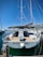 A sailboat docked at a marina, featuring a spacious deck and dual steering wheels. The sky is clear with bright blue hues, and several other boats are visible in the background. A small red flag can be seen on the boat's mast, and the marina is bustling with nautical activity.