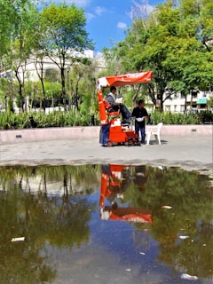 A shoe shiner is working on a red stand in an outdoor park setting. Nearby, a man is seated on a chair, waiting. The surroundings include green trees and a reflection in a large puddle of water on the ground.