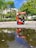 A shoe shiner is working on a red stand in an outdoor park setting. Nearby, a man is seated on a chair, waiting. The surroundings include green trees and a reflection in a large puddle of water on the ground.