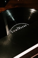 A close-up view of a vinyl record partially in its sleeve, with the label displaying the name of a music group. The surface of the disc reflects light, giving it a glossy appearance. The background is dark, contrasting with the lighter text on the label.