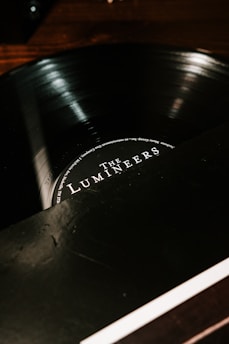 A close-up view of a vinyl record partially in its sleeve, with the label displaying the name of a music group. The surface of the disc reflects light, giving it a glossy appearance. The background is dark, contrasting with the lighter text on the label.
