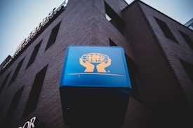 A large blue sign with a logo depicting human figures standing on an open hand is prominently displayed on the side of a brick building. The building's wall features rectangular windows and an industrial, urban appearance. The sky is overcast, creating a somber atmosphere.