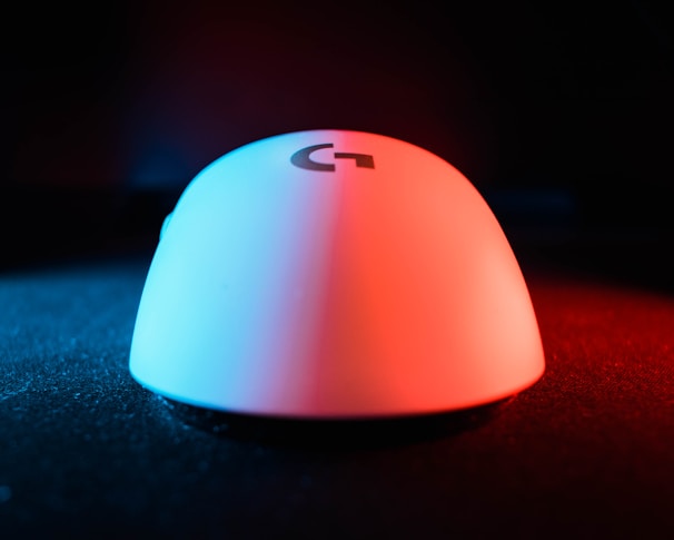 A computer mouse is illuminated with blue and red lighting, creating a striking contrast. It rests on a textured black surface, and there is a logo visible on the mouse's surface.