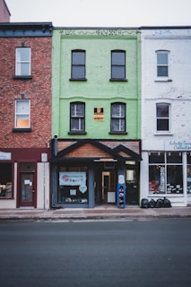 A row of three connected buildings, each with distinct architectural styles. The central building is painted bright green with a sign indicating a location for lease. It has four windows, and graffiti is visible at the top. To the left is a red brick building with two recessed windows, while the right building is white with a display window featuring a 'SALE' sign. A street runs in front of the buildings with a visible sidewalk.