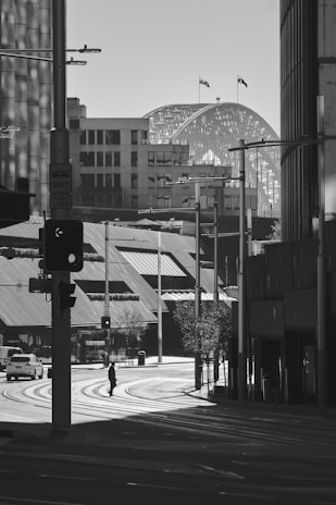 A black and white cityscape with a prominent bridge in the background. The scene includes urban buildings, a traffic light, and a pedestrian crossing an empty street in the foreground. The lighting creates dramatic shadows and highlights the architectural elements.