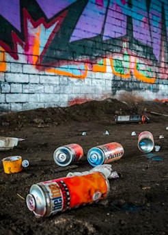 A series of spray paint cans are scattered on a dirt floor in front of a brick wall adorned with colorful graffiti. The graffiti features vibrant colors and abstract shapes, adding a lively contrast to the otherwise gritty environment.