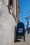 A blue United States Postal Service mailbox is positioned against a stone building with classical architectural features, such as columns and detailed moldings. The sidewalk and traffic lights are visible, along with a clear blue sky and a leafless tree in the background.