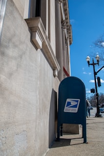 A blue United States Postal Service mailbox is positioned against a stone building with classical architectural features, such as columns and detailed moldings. The sidewalk and traffic lights are visible, along with a clear blue sky and a leafless tree in the background.