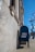 A blue United States Postal Service mailbox is positioned against a stone building with classical architectural features, such as columns and detailed moldings. The sidewalk and traffic lights are visible, along with a clear blue sky and a leafless tree in the background.