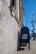A blue United States Postal Service mailbox is positioned against a stone building with classical architectural features, such as columns and detailed moldings. The sidewalk and traffic lights are visible, along with a clear blue sky and a leafless tree in the background.