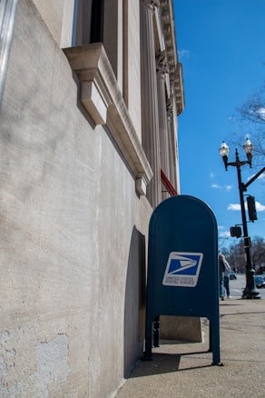 A blue United States Postal Service mailbox is positioned against a stone building with classical architectural features, such as columns and detailed moldings. The sidewalk and traffic lights are visible, along with a clear blue sky and a leafless tree in the background.