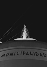 A black and white photograph of a building with the word 'Municipalidad' displayed prominently near the bottom. The top part of the building features an architectural structure with lines extending upward toward the dark sky.