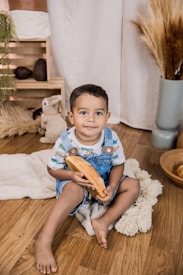 A young child is seated on a wooden floor, holding a piece of bread. The child is wearing a striped shirt and denim overalls. Behind them are decorative elements such as a soft blanket, a plush toy, dried plants in a vase, and wooden decorative items. The room has a cozy and rustic atmosphere.