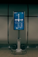A digital display screen mounted on a metal stand shows a map of gates at an airport. The screen is located in a dimly lit area with tiled flooring and a metallic, industrial-looking backdrop. The map includes labels for gates ranging from 30 to 59 and features various icons to indicate facilities like cafes and shops.