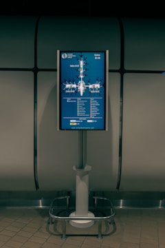 A digital display screen mounted on a metal stand shows a map of gates at an airport. The screen is located in a dimly lit area with tiled flooring and a metallic, industrial-looking backdrop. The map includes labels for gates ranging from 30 to 59 and features various icons to indicate facilities like cafes and shops.
