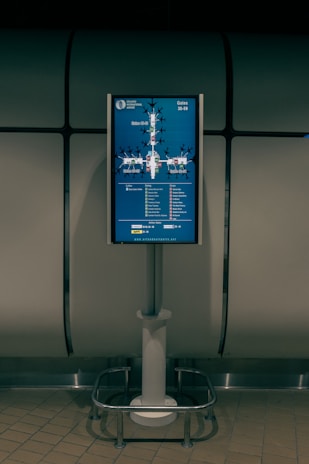 A digital display screen mounted on a metal stand shows a map of gates at an airport. The screen is located in a dimly lit area with tiled flooring and a metallic, industrial-looking backdrop. The map includes labels for gates ranging from 30 to 59 and features various icons to indicate facilities like cafes and shops.