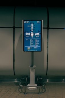 A digital display screen mounted on a metal stand shows a map of gates at an airport. The screen is located in a dimly lit area with tiled flooring and a metallic, industrial-looking backdrop. The map includes labels for gates ranging from 30 to 59 and features various icons to indicate facilities like cafes and shops.