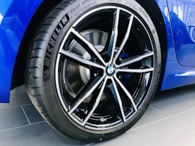 A detailed focus on the wheel of a car showcasing a Michelin tire and a stylish BMW alloy rim. The wheel is clean and glossy, with a visible BMW logo in the center of the rim. The car's body is a vibrant blue, adding to the sleek and modern appearance against the dark asphalt ground.