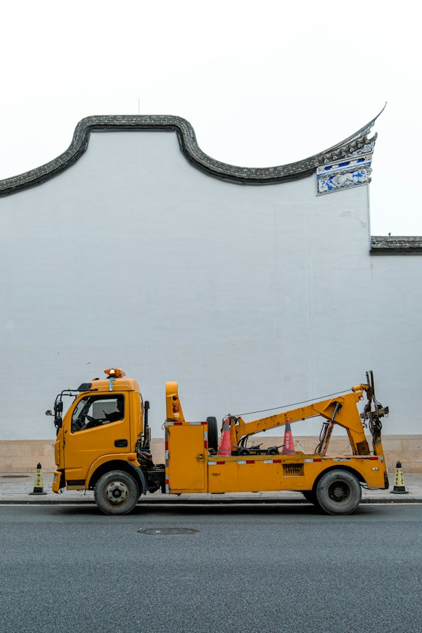 Yellow crane-style tow truck parked along a street beside an ornate white wall