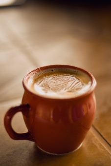 A warm ceramic mug filled with creamy coffee, placed on a smooth, wooden surface. The frothy surface of the coffee shows a detailed, appealing texture. Soft lighting creates a cozy and inviting atmosphere.