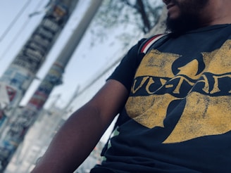 A person wearing a black T-shirt featuring a distinctive yellow design, which seems to be a logo. The background includes a blurry view of utility poles and a wire fence, suggesting an urban setting.