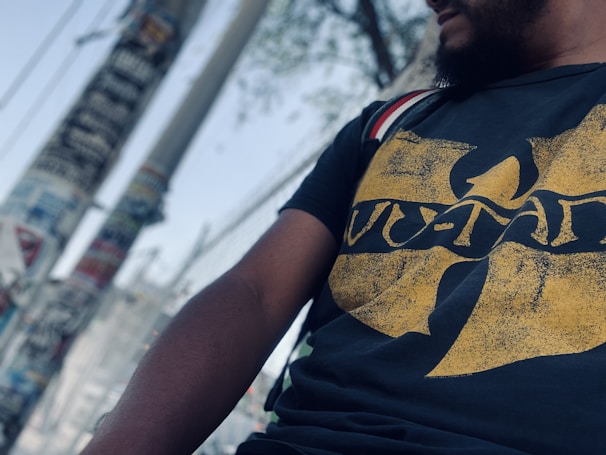 A person wearing a black T-shirt featuring a distinctive yellow design, which seems to be a logo. The background includes a blurry view of utility poles and a wire fence, suggesting an urban setting.