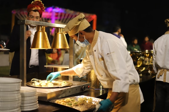 A chef wearing a white uniform and a beige hat is preparing food under warm lighting. He is using blue gloves and wearing a face mask while standing at a serving station. In the background, a man in a formal suit is viewing the preparation, and there are various cooking utensils and serving dishes visible.