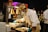 A chef wearing a white uniform and a beige hat is preparing food under warm lighting. He is using blue gloves and wearing a face mask while standing at a serving station. In the background, a man in a formal suit is viewing the preparation, and there are various cooking utensils and serving dishes visible.