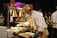 A chef wearing a white uniform and a beige hat is preparing food under warm lighting. He is using blue gloves and wearing a face mask while standing at a serving station. In the background, a man in a formal suit is viewing the preparation, and there are various cooking utensils and serving dishes visible.