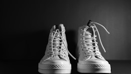 A pair of white high-top sneakers is positioned side by side against a dark background. The sneakers have visible laces and textured soles, creating a contrast against the simplicity of the setting.