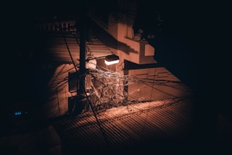 A tangled mass of electrical wires hangs near a utility pole, dimly illuminated by a single streetlight. The scene is set against an urban backdrop with visible shadows and contrasting light, suggesting a disorganized network of overhead cables.