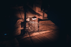 A tangled mass of electrical wires hangs near a utility pole, dimly illuminated by a single streetlight. The scene is set against an urban backdrop with visible shadows and contrasting light, suggesting a disorganized network of overhead cables.