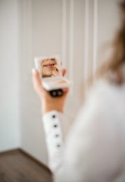 A person holds a compact mirror in one hand while applying lipstick with the other. The focus is on the lips and the lipstick application, captured in the reflection of the mirror. The scene is softly lit, creating a tranquil and intimate atmosphere.
