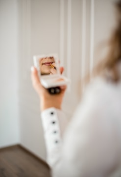 A person holds a compact mirror in one hand while applying lipstick with the other. The focus is on the lips and the lipstick application, captured in the reflection of the mirror. The scene is softly lit, creating a tranquil and intimate atmosphere.