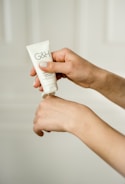 A person's hands are applying cream from a tube labeled 'G&H' onto the back of one hand. The background is soft and neutral, creating a minimalistic look.