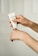A person's hands are applying cream from a tube labeled 'G&H' onto the back of one hand. The background is soft and neutral, creating a minimalistic look.