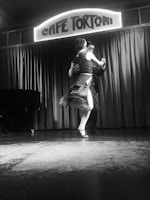 A couple performs a dance on a stage, with a vintage-style sign above them reading 'Café Tortoni'. The dancers are captured mid-motion, indicating a sense of fluidity and grace. The setting includes a closed curtain backdrop and a piano visible on the left-hand side.