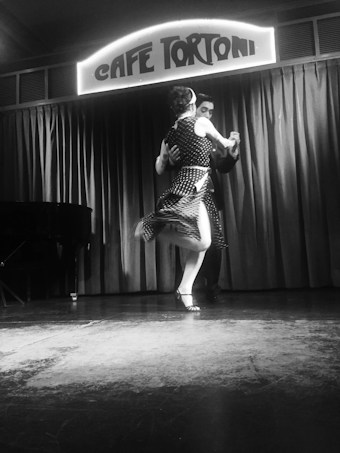 A couple performs a dance on a stage, with a vintage-style sign above them reading 'Caf&eacute; Tortoni'. The dancers are captured mid-motion, indicating a sense of fluidity and grace. The setting includes a closed curtain backdrop and a piano visible on the left-hand side.
